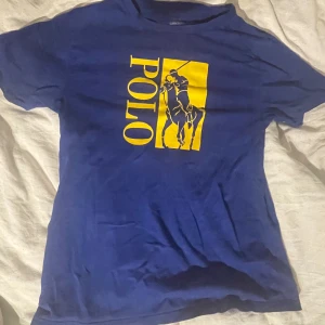 Blå t-shirt från Ralph Lauren (160 cm) - Säljer en blå t-shirt från Ralph Lauren med ett gult Polo-tryck på framsidan. T-shirten är kortärmad och perfekt för en avslappnad stil. Klassisk design med en modern touch. Storlek 160 cm