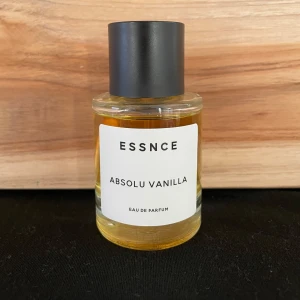 Essnce Absolu Vanilla Eau de Parfum - Säljer en flaska Essnce Absolu Vanilla Eau de Parfum. Flaskan är genomskinlig med en svart kork och innehåller en gyllene doftvätska. Den har en enkel och stilren design med en vit etikett.