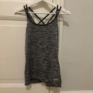 Grå träningslinne från Nike - Säljer ett grått träningslinne från Nike med Dri-FIT-teknologi. Linnet har ett melerat mönster och dubbla korsade axelband för extra stöd. Perfekt för träning och aktivitet.