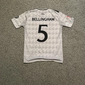 Vit Real Madrid fotbollströja - Snygg vit Real Madrid fotbollströja från Adidas med svart text och logga. Tröjan har korta ärmar och ett subtilt mönster. Perfekt för fans som vill visa sitt stöd.