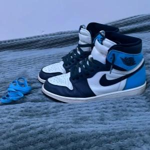 Nike Air Jordans 1 unc retro high obsidian storlek 41 - Snygga Nike Air Jordans i blått och vitt med svarta detaljer. Skorna har klassisk snörning och en hög siluett. De är i storlek 41 . Extra blåa skosnören medföljer. Har en liten defekt på vänstra skon som går o ses på bild men är knappt märkbar. kontakta för mer info! Vill rensa min garderob och om det kommer snabbt svar kan man få det för billigare