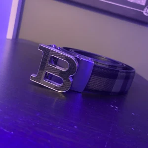 Burberry bälte  - Snyggt bälte med ett stort B-format spänne i silver. Bältet har ett stilrent rutigt mönster i svart och grått, perfekt för att ge en extra touch till din outfit. Tillverkat i ett robust material som ger en lyxig känsla.