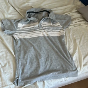 Grå och vit randig pikétröja från Calvin Klein - Snygg grå pikétröja från Calvin Klein med vita ränder över bröstet. Tröjan har en klassisk krage och korta ärmar, perfekt för en avslappnad stil. Märkesloggan är diskret broderad på bröstet.