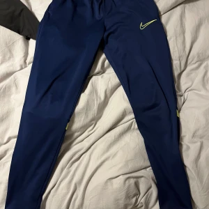 Blå träningsbyxor från Nike - Snygga blå träningsbyxor från Nike med neongula detaljer. Perfekta för träning med en bekväm passform och elastisk midja. Byxorna har en smal design och är gjorda i ett lätt och flexibelt material.