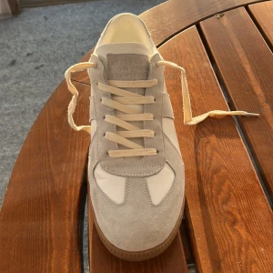 Vita sneakers från Maison Margiela - Snygga vita sneakers från Maison Margiela med detaljer i beige mocka och en gummisula. Skorna har klassisk snörning och en stilren design som passar perfekt för en trendig look. Dust bag och låda följer givetvis också med!
