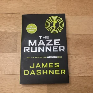 The Maze Runner - Säljer boken 'The Maze Runner' av James Dashner på engelska. Det är den första boken i den populära Maze Runner-serien. Omslaget är svart med vit och grön text samt en cirkulär labyrintdesign. Perfekt för dig som gillar spännande och dystopiska berättelser.