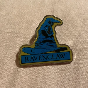 Ravenclaw pin - Snygg pin med Ravenclaw-tema från Harry Potter. Designen visar en blå hatt med texten 'Ravenclaw' i svart. 