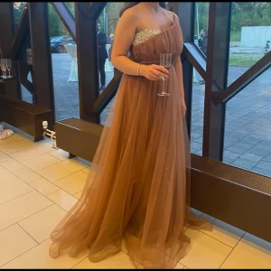 Brun axelbandslös balklänning - Elegant brun axelbandslös balklänning med glittriga detaljer på bysten. Klänningen har en lång, flödande kjol i ett lätt och luftigt material som ger en drömlik känsla. Perfekt för en speciell kväll!