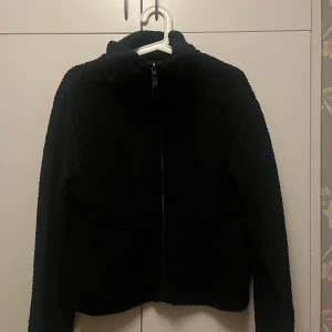 Svart teddyjacka från H&M - Mysig svart teddyjacka från H&M med dragkedja framtill. Perfekt för kyliga dagar med sin fluffiga och varma design. Jackan har långa ärmar och en bekväm passform.