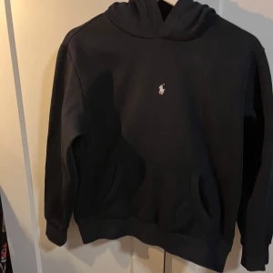 Svart hoodie från Ralph Lauren - Säljer en stilren svart hoodie från Ralph Lauren med det klassiska logotypbroderiet på bröstet. Tröjan har en bekväm passform och är perfekt för en avslappnad look. Den har långa ärmar och en praktisk huva. Köpte hoodie inför 1200 ungefär så säljer den för ett jättebra pris.