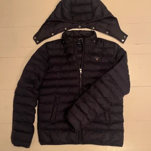 Gant Dunjacka - Säljer en stilren svart dunjacka från GANT med avtagbar huva och dragkedja framtill. Sorleken passar Small. Skriv vid minsta fundering👍