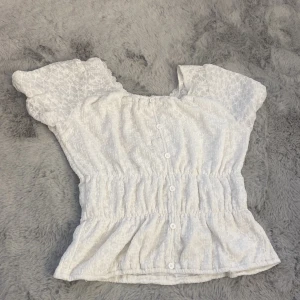 Vit blus från ONLY - Säljer en söt vit blus från ONLY. Den har korta ärmar och ett elastiskt midjeband som ger en fin passform. Blusen är dekorerad med små knappar framtill och har ett diskret blommigt mönster. Perfekt för vår och sommar!