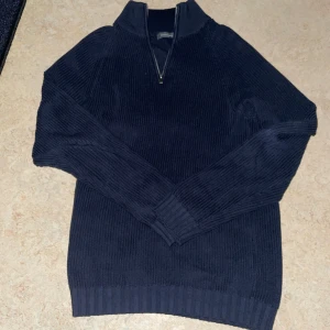 Mörkblå stickad tröja med dragkedja - Säljer en snygg mörkblå stickad tröja med dragkedja vid halsen. Perfekt för kyliga dagar och passar både till vardags och lite finare tillfällen. Tröjan har långa ärmar och ribbade muddar vid ärmslut och nederkant. Supermysig och stilren!