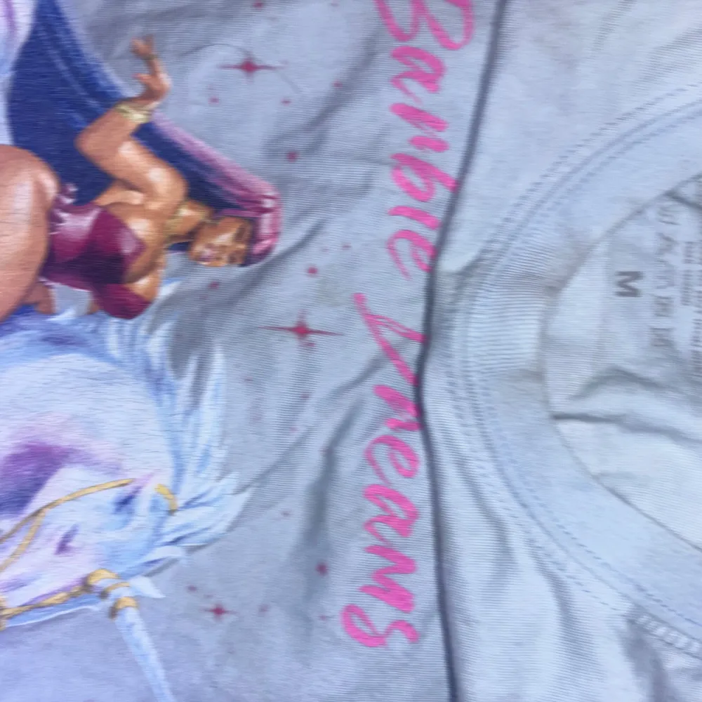 Säljer en t-shirt med ett coolt Barbie Dreams-tryck på framsidan. Den har en färgglad illustration av en kvinna på en enhörning och texten 'Barbie Dreams'. På baksidan finns texten 'Nicki World Tour’ med en lista över städer hon besökte på touren2019. T-shirts.