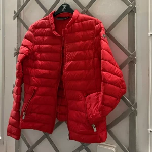 Jacka peak performance - En röd jacka med en dragkedja framtill och två fickor med dragkedja på sidorna. Jackan har en hög krage och en liten logga på vänster bröst. Den är använd fåtal gånger!