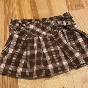Brun och rosa rutig kjol med spännen - Säljer en superfin brun och rosa rutig kjol från Kids by Lindex i storlek 140. Kjolen har coola spännen på sidorna som ger en extra detalj. Perfekt för höst och vår, och funkar både till vardags och fest. 🩷🤎