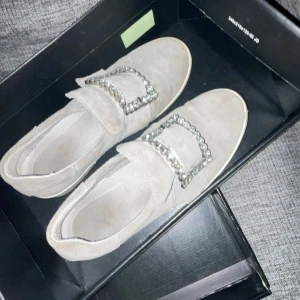 Beige mocka loafers med strassdetaljer - Säljer ett par snygga beige loafers i mocka med glittrande strassdetaljer på ovansidan. Perfekta för att ge en touch av glamour till vardagsoutfiten. De är i bra skick och kommer i originalkartong.