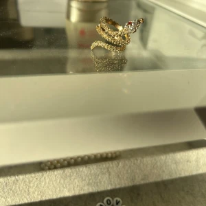Guldfärgad ring med glittriga stenar - Säljer en superfin guldfärgad ring som är formad som en orm och dekorerad med glittriga stenar. Den har en cool design med en röd sten som öga, vilket ger den en unik look. Perfekt för att piffa upp vilken outfit som helst!