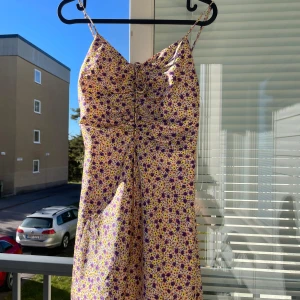 Blommig sommarklänning - Säljer en superfin blommig sommarklänning i ljusa färger med lila och gula blommor. Klänningen är ärmlös och har smala axelband, perfekt för varma sommardagar. Den har en söt knytning framtill som ger en extra touch. Klänningen är kort och passar perfekt för en dag på stranden eller en sommarfest. 🌸