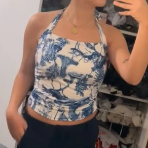 Vit och blå halterneck topp - Säljer en snygg vit halterneck topp med blått blommönster. Toppen har en åtsittande passform och är perfekt för sommardagar. Den har en stilren design med knytning i nacken.