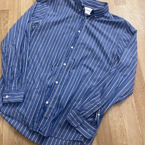 Blå randig skjorta från These Glory Days - Snygg blå skjorta med vita ränder från These Glory Days. Skjortan har långa ärmar och knappar framtill. Perfekt för en stilren look.