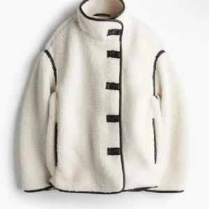 Vit teddyjacka med svarta detaljer - Säljer en supermysig vit teddyjacka med svarta kantdetaljer och knäppning framtill. Jackan har en hög krage och långa ärmar, perfekt för kyliga dagar. Den är både stilren och bekväm, ett måste i garderoben!