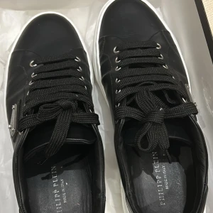 Svarta sneakers från Philipp Plein - Snygga svarta sneakers från Philipp Plein med klassisk snörning och vit sula. Tillverkade i skinn för en lyxig känsla. Perfekta för en stilren look. Dessutom bara använd bara 1 gång.