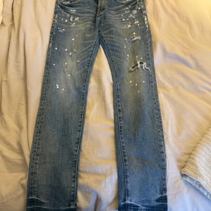 Blå jeans med slitna detaljer - Snygga blå jeans med coola slitna detaljer och färgstänk. Perfekta för en avslappnad stil. De har en klassisk femficksdesign och en bekväm passform.