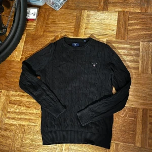 Svart kabelstickad tröja från Gant - Snygg svart kabelstickad tröja från Gant med rund halsringning. Tröjan har långa ärmar och ribbade muddar vid ärmslut och nederkant. Perfekt för en stilren look.