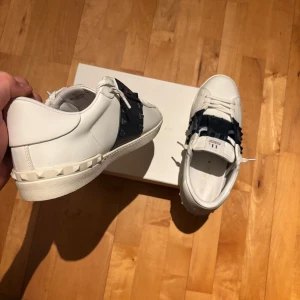 Valentino Garavanis - Snygga vita Valentino Garavani sneakers med en bred mörkblå rem över snörningen. Skorna har en stilren design med dekorativa nitar runt hälen och en bekväm sula. Perfekta för en trendig look. Skorna är köpta direkt av Valentinos hemsida vid nypris c.a 8000 kr, jag säljer dessa eftersom att de va lite för små för mig. Svinfeta skor vid svinbra pris hör av er vetja 🤙