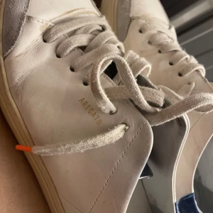 Axel Arigato skor - Snygga vita sneakers från Axel Arigato med grå och blå detaljer. Skorna har snörning och en robust sula med märkets logga. Perfekta för en stilren look.