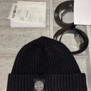 Svart ribbad mössa från Stone Island - Säljer en snygg svart ribbad mössa från Stone Island. Den är i mycket bra skick och har en stilren design med en broderad logga framtill. Perfekt för höst och vinter för att hålla dig varm och trendig. Passar de flesta tack vare sin flexibla passform.