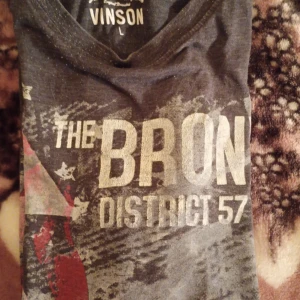 Grå t-shirt från Vinson - Säljer en grå t-shirt från Vinson med tryck 'The Bronx District 57' i vitt och rött. T-shirten har en cool urban stil med korta ärmar och rund hals. Perfekt för en avslappnad look.