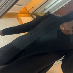 Svart kappa med bälte och huva  - Snygg svart kappa med bälte som ger en elegant look. Perfekt för kyligare dagar med sin långa design och stilrena knappar. Jätte varm och är som ny från Boozt. Inte köpt för länge sen. Köpte för 800.