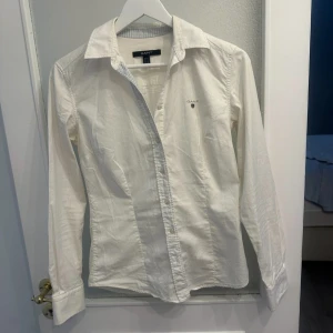 Vit långärmad skjorta från GANT - Snygg vit skjorta från GANT med broderad logga på bröstet. Skjortan har en klassisk krage och långa ärmar. Perfekt för en stilren look.