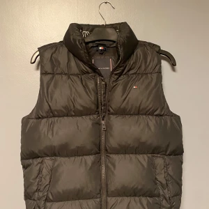 Svart dunväst från Tommy Hilfiger i storlek 164 - Snygg svart dunväst från Tommy Hilfiger med dragkedja framtill. Perfekt för kyligare dagar och ger en stilren look. Västen har en hög krage och är quiltad för extra värme.  Den är för barn i storlek i 164 