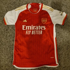 Röd Arsenal fotbollströja - Snygg röd Arsenal fotbollströja från Adidas med vita och guld detaljer. Tröjan har korta ärmar och ett subtilt randigt mönster. Perfekt för alla Arsenal-fans!