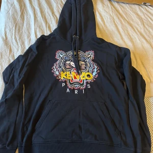 Svart hoodie från Kenzo - Säljer en svart hoodie från Kenzo med ett färgglatt tigerbroderi på framsidan. Tröjan har en klassisk känguruficka och justerbar huva med dragsko. Finns en fel flaw, litet hål på armen samt vid fickan i hörnen.