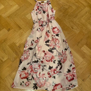 Blommig långklänning från Lily Bleu - Säljer en vacker långklänning från Lily Bleu med ett blommigt mönster i rosa och svart på en ljus bakgrund. Klänningen är ärmlös och tillverkad i 100% polyester, perfekt för sommarens festligheter. 🌸