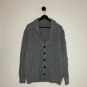 100% Kashmir Cardigan  - Säljer en mysig grå stickad kofta med kabelstickat mönster och svarta knappar. Koftan har en bred krage och långa ärmar, perfekt för kyliga dagar. Den är både stilren och bekväm. Den är i 100% Kashmir vilket gör den extremt skön! Hör gärna av er vid flera frågor eller funderingar! 