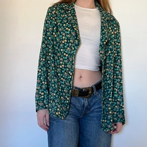 Vintage Blommig kavaj i grönt - Säljer en snygg grön tunn kavaj med blommigt mönster. Kavajen har långa ärmar och knappar framtill. Perfekt för att ge en färgglad touch till din outfit. Passar bra till jeans eller kjol. #vintage 