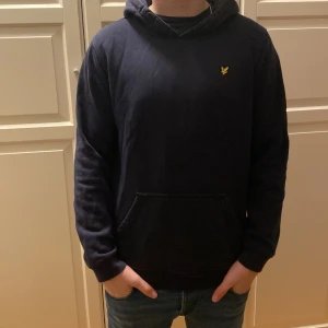 Mörkblå hoodie med gul logga - Säljer en stilren mörkblå hoodie från Lyle & Scott med en gul logga på bröstet. Tröjan har en klassisk känguruficka och en bekväm huva. Perfekt för en avslappnad stil.
