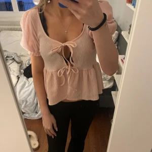 Rosa blus med knytning - Söt rosa blus med korta puffärmar och knytning framtill. Den har en lätt volangdetalj vid midjan som ger en feminin touch. Perfekt för en avslappnad stil. Från ginatricot 