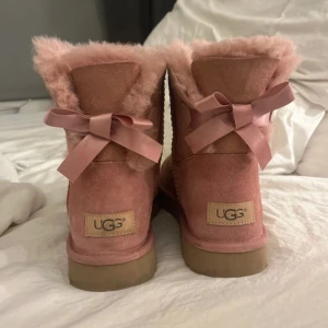 Rosa UGG boots med rosett - Säljer ett par rosa UGG boots i mocka med mjukt foder och dekorativa rosetter baktill. Perfekta för att hålla fötterna varma och stiliga under kyliga dagar. svåra att få tag i köptes för över 3000 och de är aldrig använda