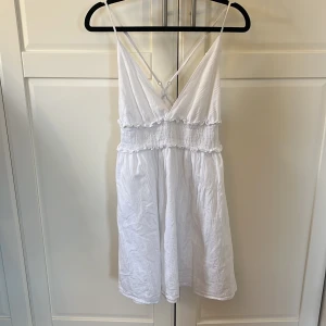 Vit sommarklänning med smala axelband - Säljer en söt vit klänning med smala axelband och v-ringad design. Klänningen har en elastisk midja med volangdetaljer och är perfekt för varma sommardagar. Den är kort och luftig, vilket gör den idealisk för en avslappnad stil.