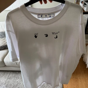 Vit t-shirt med tryck från Off-White - Säljer en vit t-shirt från Off-White med ett unikt tryck i blå och gröna nyanser på baksidan. Framsidan har en minimalistisk design med Off-White-logotypen. Perfekt för en stilren och modern look.