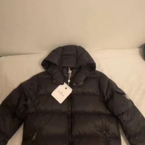 Moncler Maya - Säljer en snygg svart dunjacka från Moncler med huva och dragkedja. Jackan har en Moncler-logga på ärmen och praktiska fickor med knappar. Perfekt för kyliga dagar! Den är aldrig använt i storlek s säljer den på grund av i fel storlek för mig!
