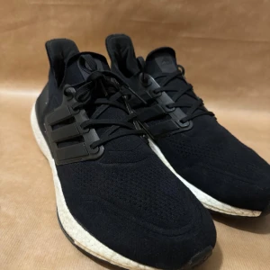 Svarta Adidas Ultraboost sneakers - Snygga svarta Adidas Ultraboost sneakers med stickad ovandel och klassisk Boost-sula för extra komfort. Skorna har snörning och de ikoniska tre ränderna på sidan. Perfekta för både träning och vardag. Storlek 46 1/3 