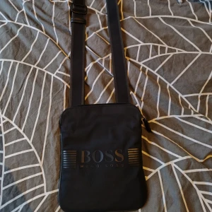 Svart axelväska från Hugo Boss - Snygg svart axelväska från Hugo Boss med justerbar rem och dragkedja. Perfekt för att bära dina nödvändigheter med stil. Väskan har en stilren design med BOSS-logga på framsidan.