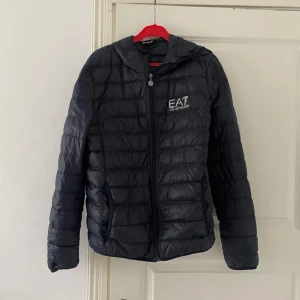 Svart dunjacka från EA7 - Säljer en stilren svart dunjacka från EA7. Jackan har en quiltad design med dragkedja framtill och en praktisk huva. Perfekt för kyligare dagar och ger en sportig look. Passar bra till både vardag och mer avslappnade tillfällen.